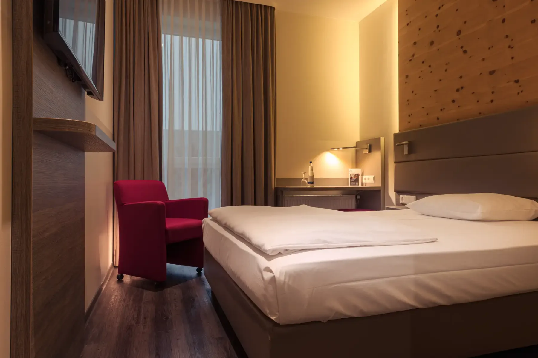 Habitación de hotel con una cama doble hecha, un sillón rojo, un televisor montado y una iluminación cálida.