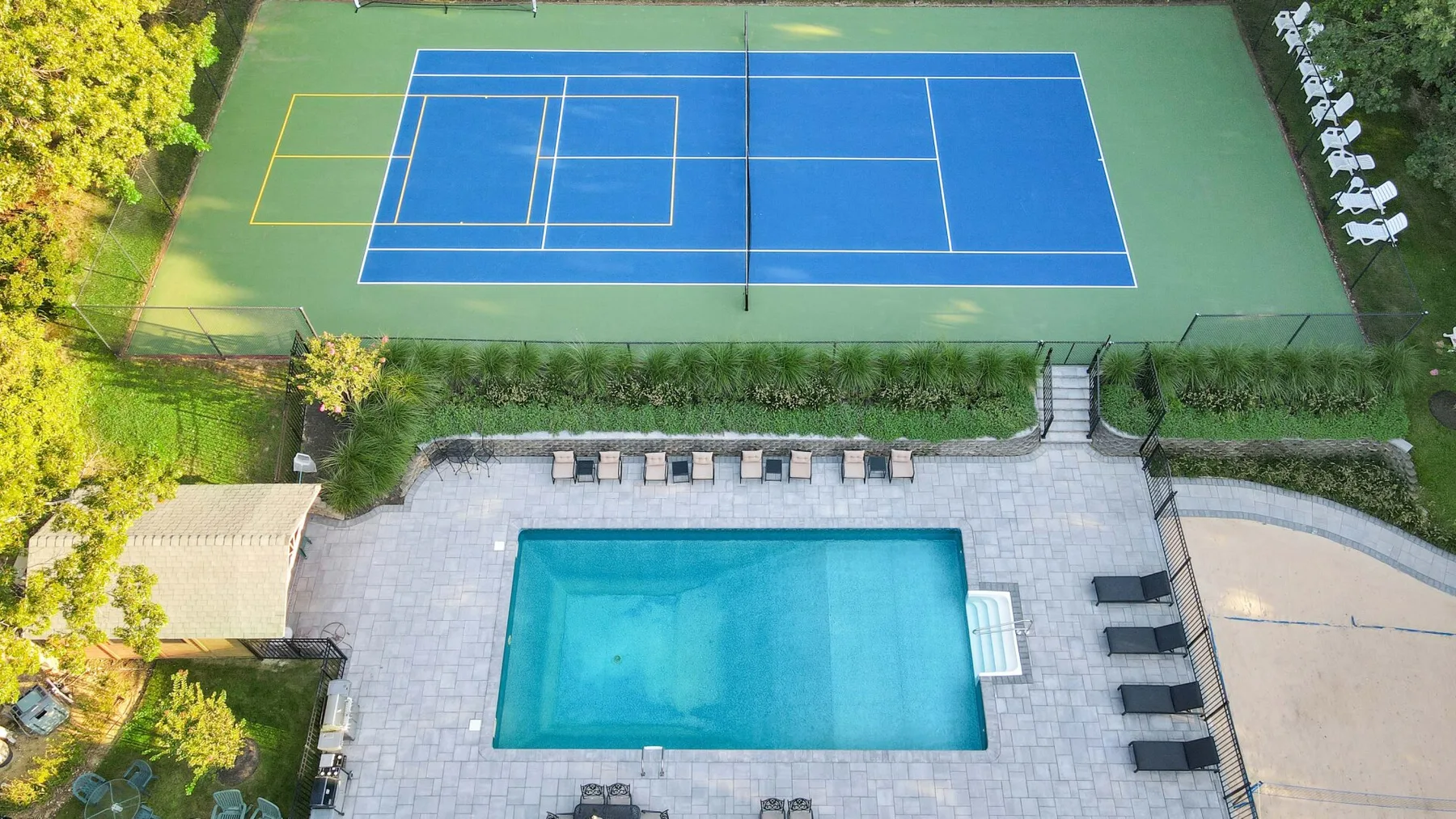 An outdoor pool and blue tennis court are surrounded by paved areas and greenery.