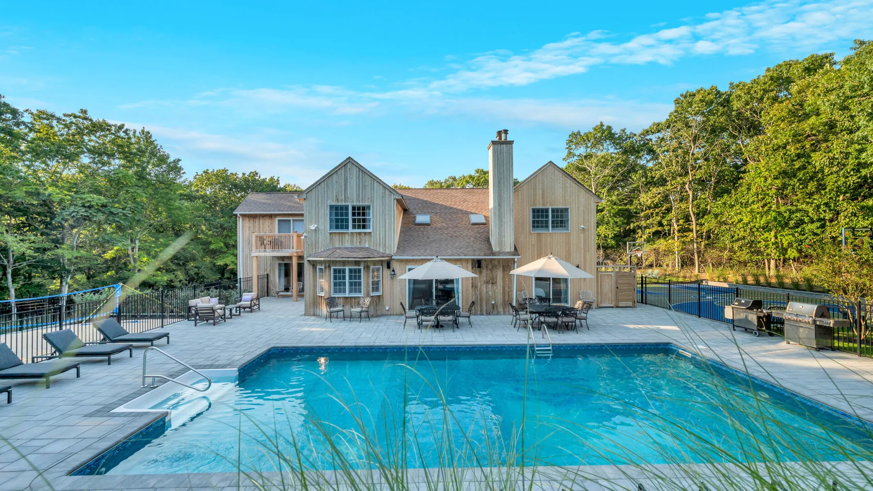 A large outdoor swimming pool sits beside a wood-sided house and patio seating.