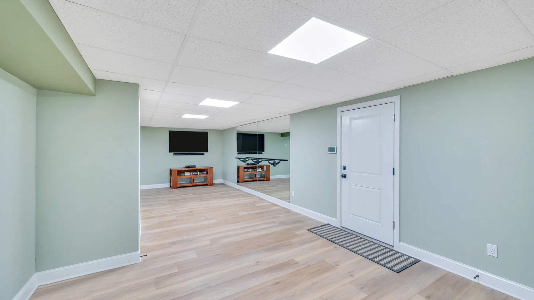 A finished basement room with light flooring, wall-mounted screens, and a closed door.