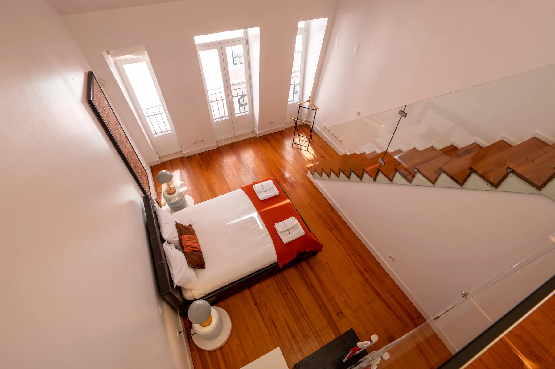 A bedroom with a bed beside tall windows and a wooden staircase.