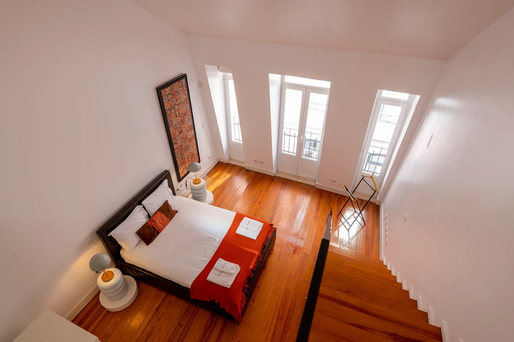 A bedroom with a bed beside tall windows and a wooden staircase.
