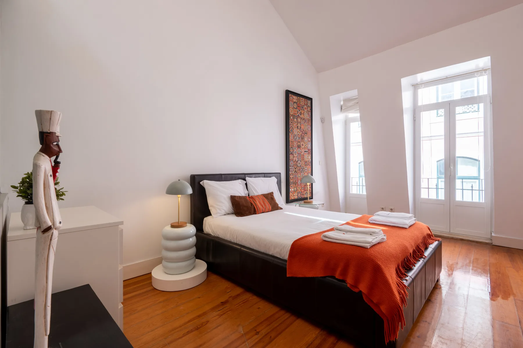 A bedroom with a bed dressed in white and orange linens beside tall windows.