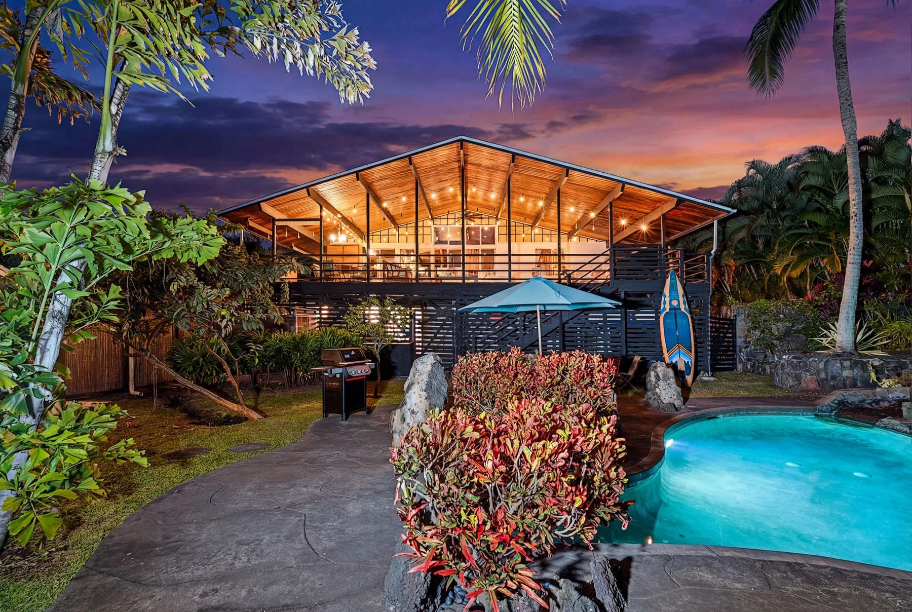 An illuminated open-sided pavilion stands beside a swimming pool at dusk.