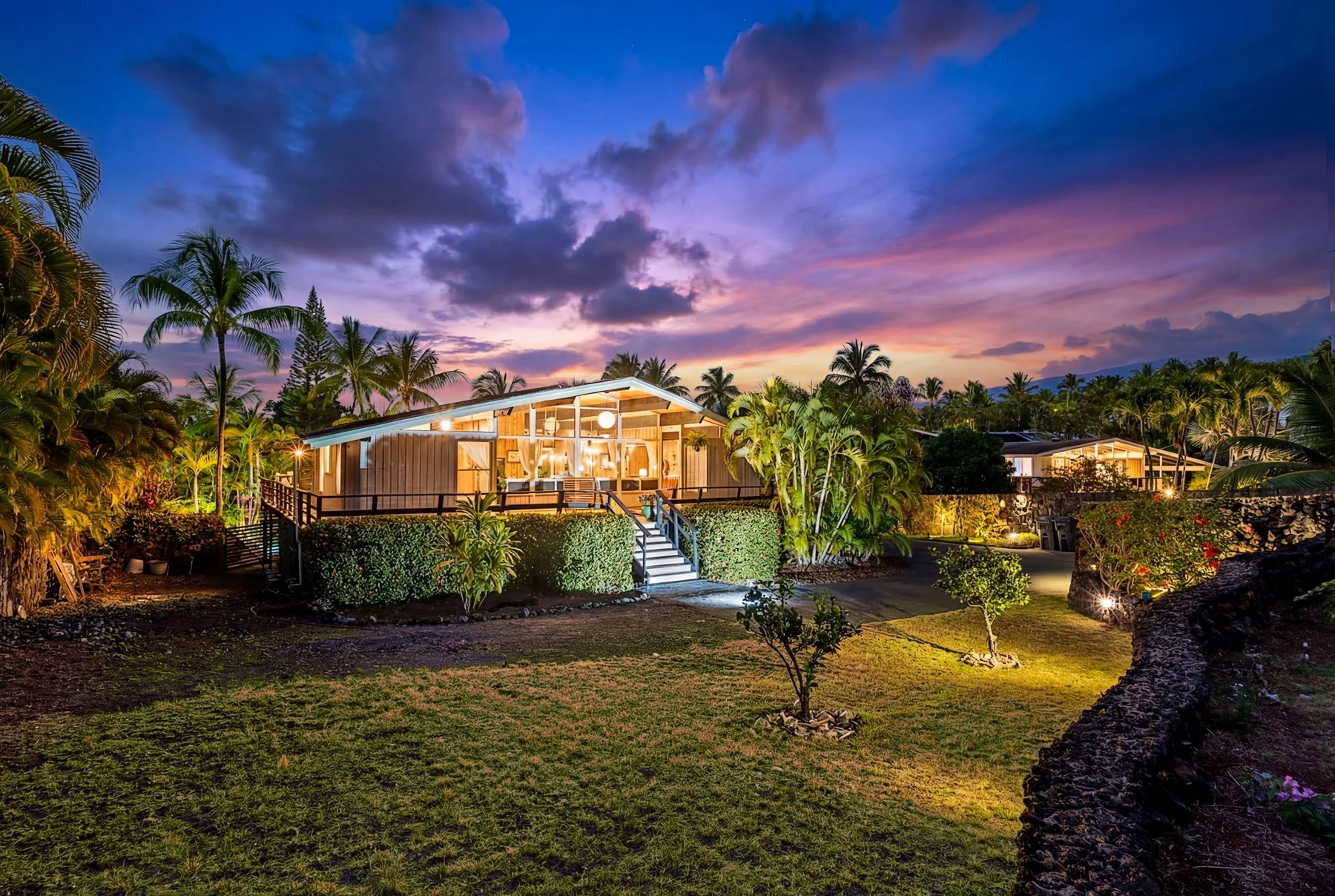 A warmly lit house stands among palm trees beneath a colorful sunset sky.