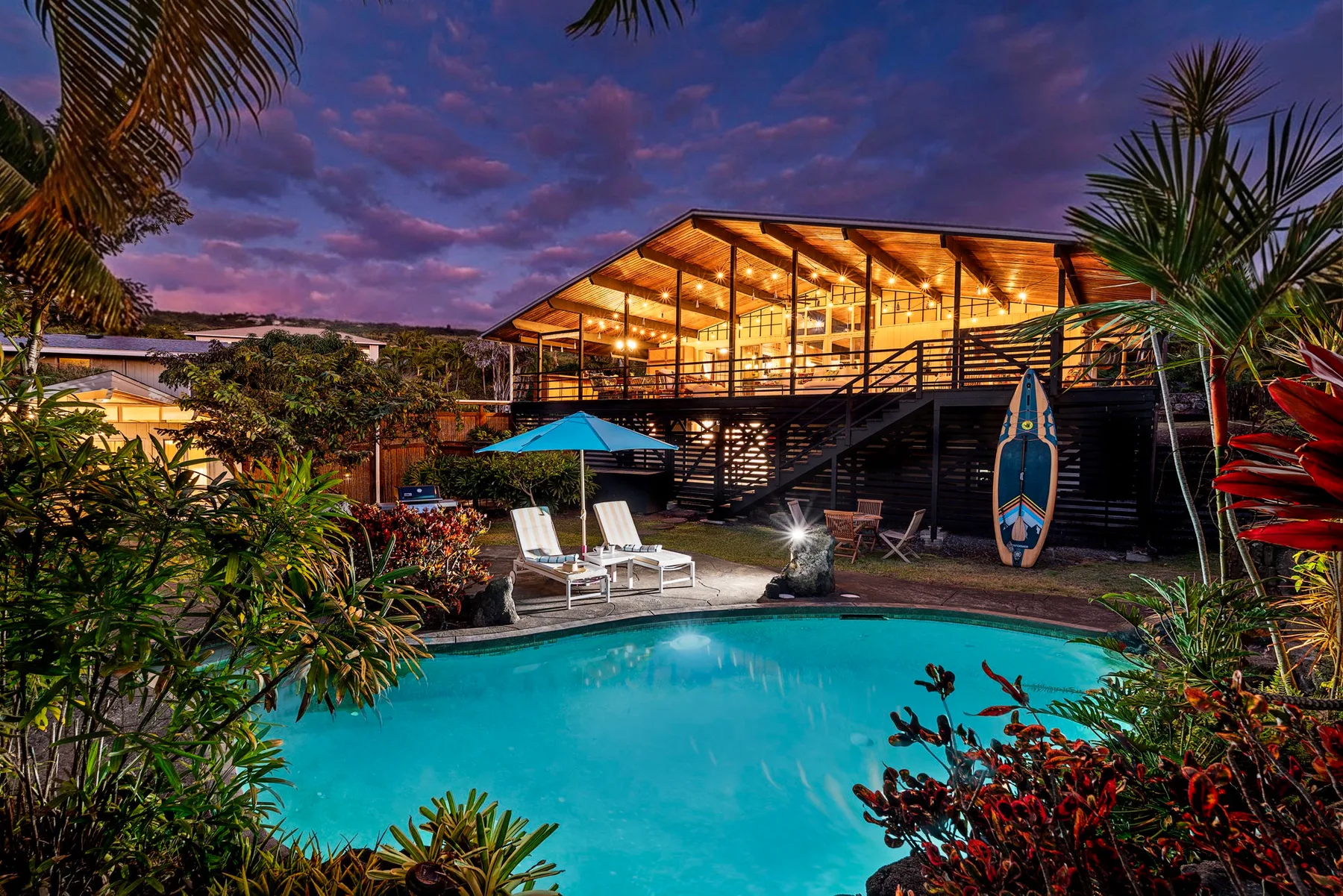 An outdoor swimming pool sits beside a warmly lit building and tropical plants at dusk.
