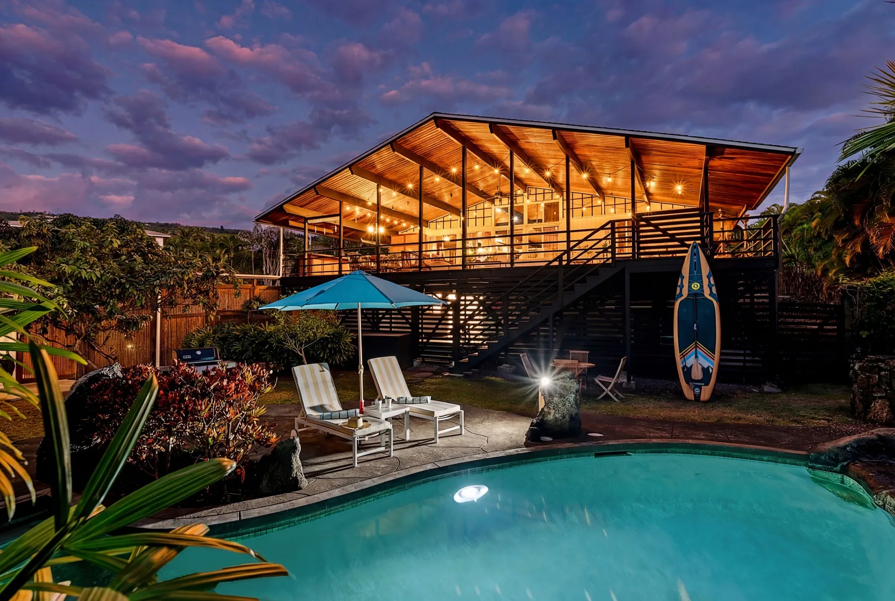 An outdoor pool sits in front of a raised, warmly lit building at dusk.