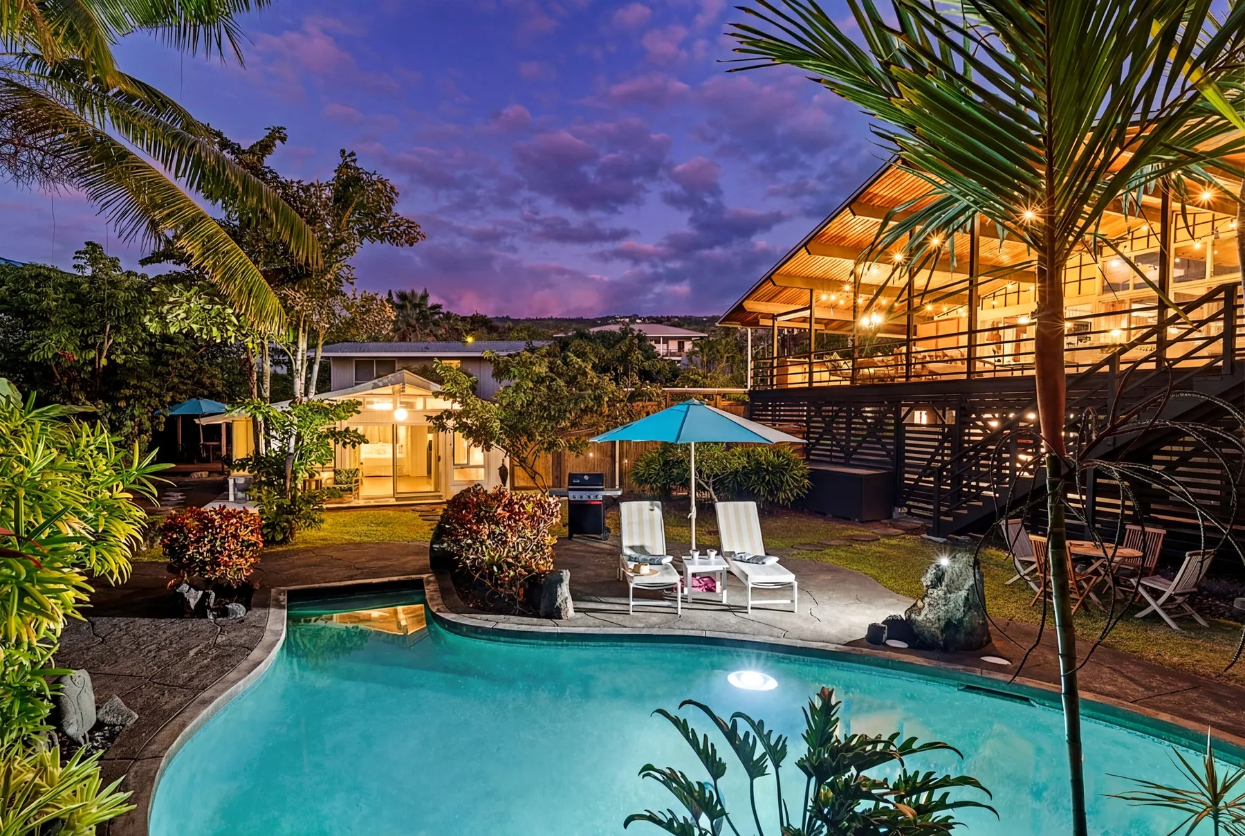 An outdoor pool beside a brightly lit building, with palm trees and a patio umbrella.