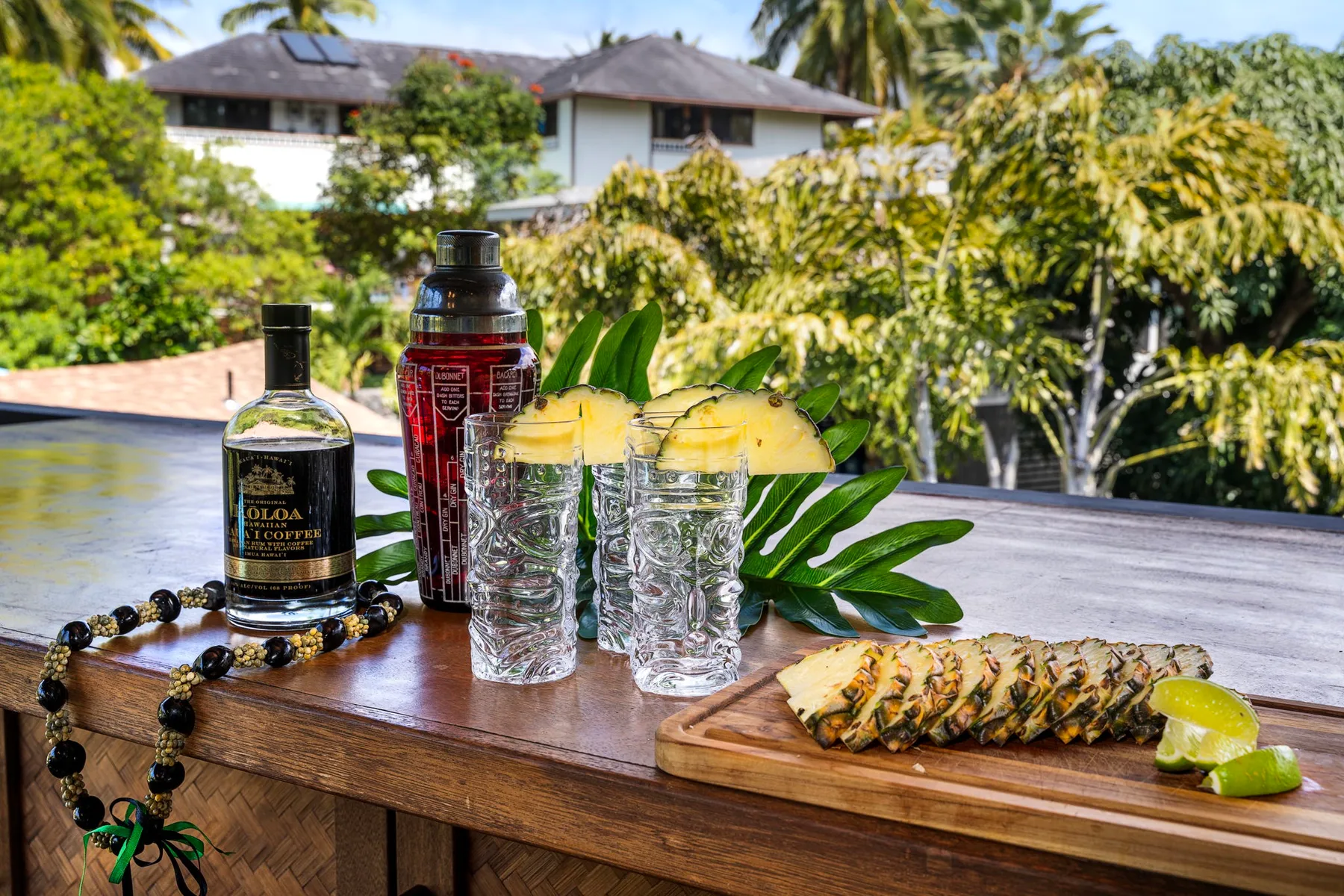 A wooden outdoor table displays drinks, fruit, and a sliced pineapple amid tropical greenery.