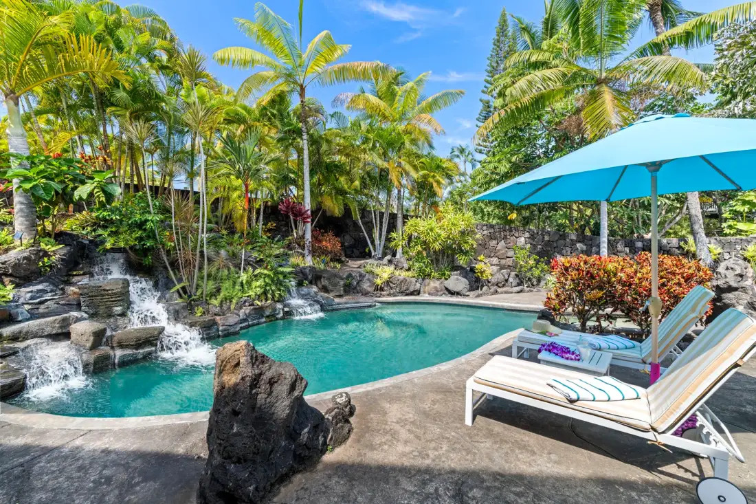A tropical outdoor pool with a small waterfall and lounge seating under an umbrella.