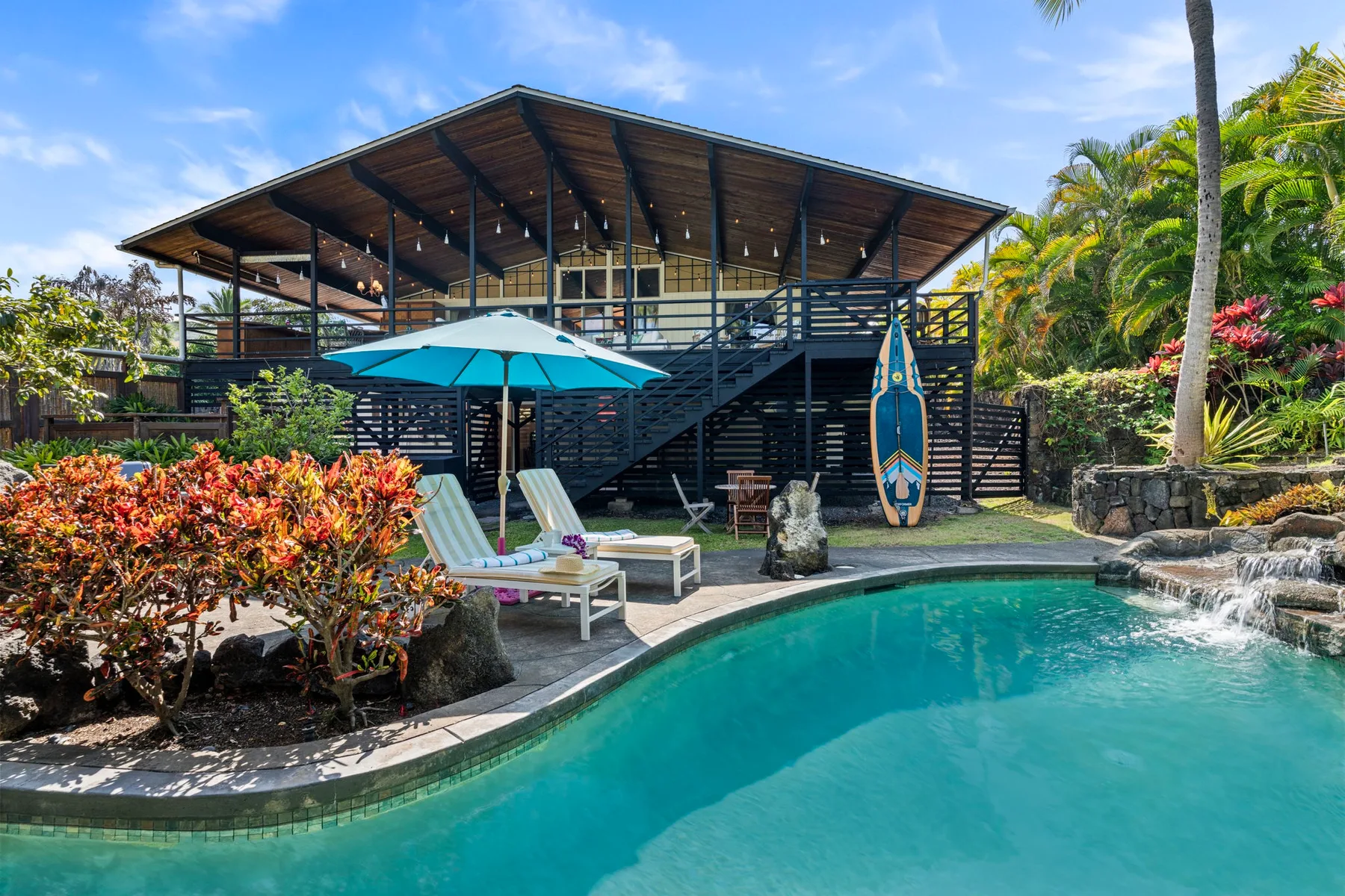 A tropical outdoor pool sits beside a raised wooden building and lounge chairs.