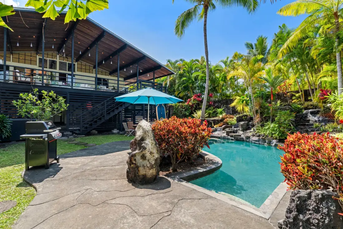 A tropical outdoor pool beside a raised building, with a grill on the patio.