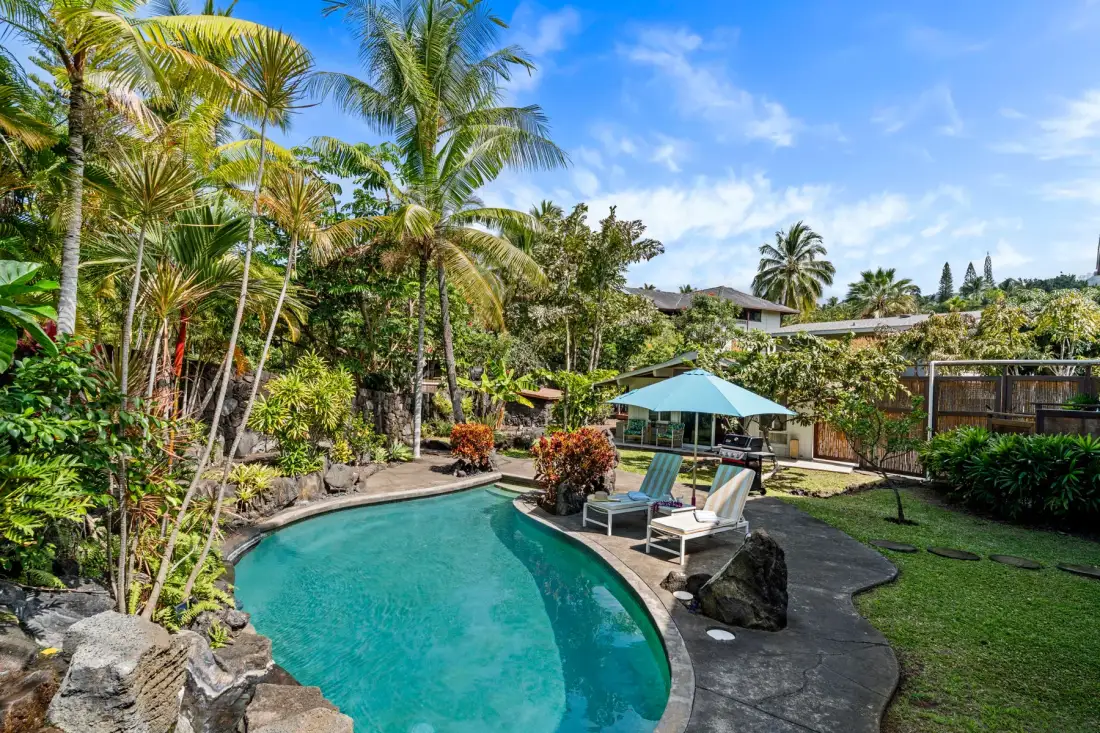 A garden swimming pool sits among tropical plants, with shaded seating beside the water.
