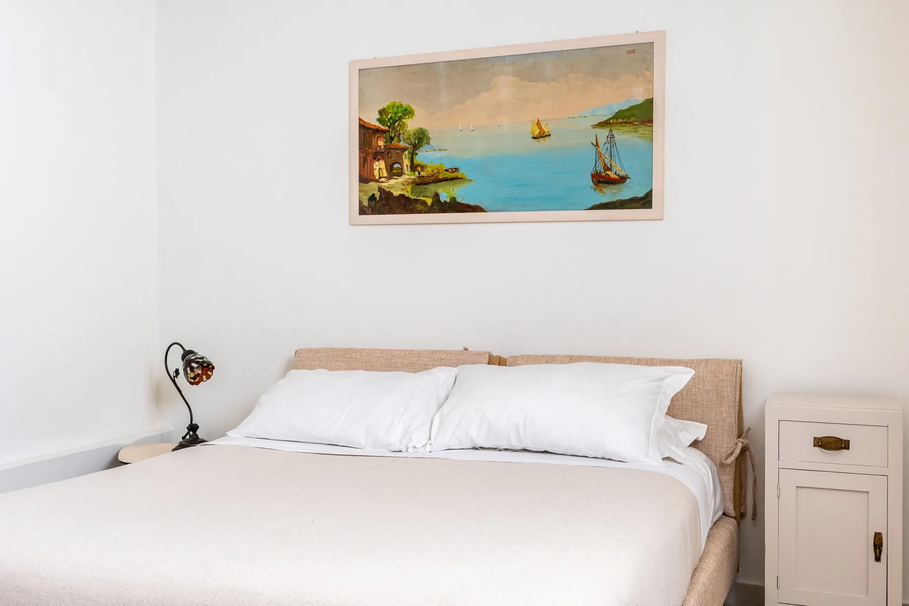 Un dormitorio con una cama bien tendida y un cuadro enmarcado de una escena costera.