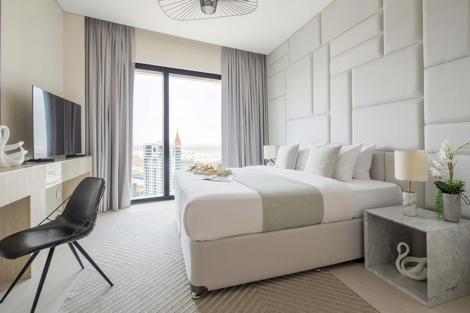 Un dormitorio luminoso con una cama bien tendida junto a ventanales y cortinas grises.