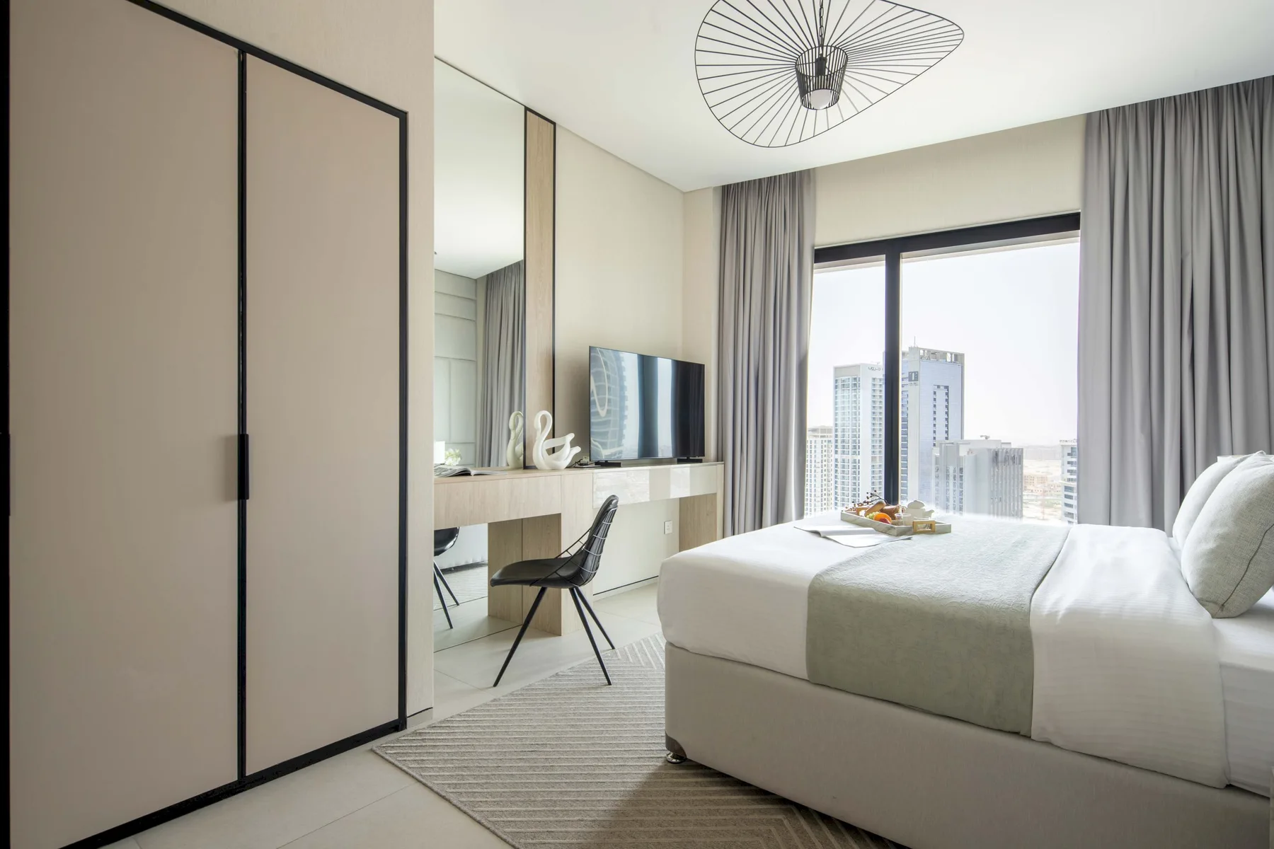 Un dormitorio con cama, escritorio y grandes ventanas con vistas a edificios de la ciudad.