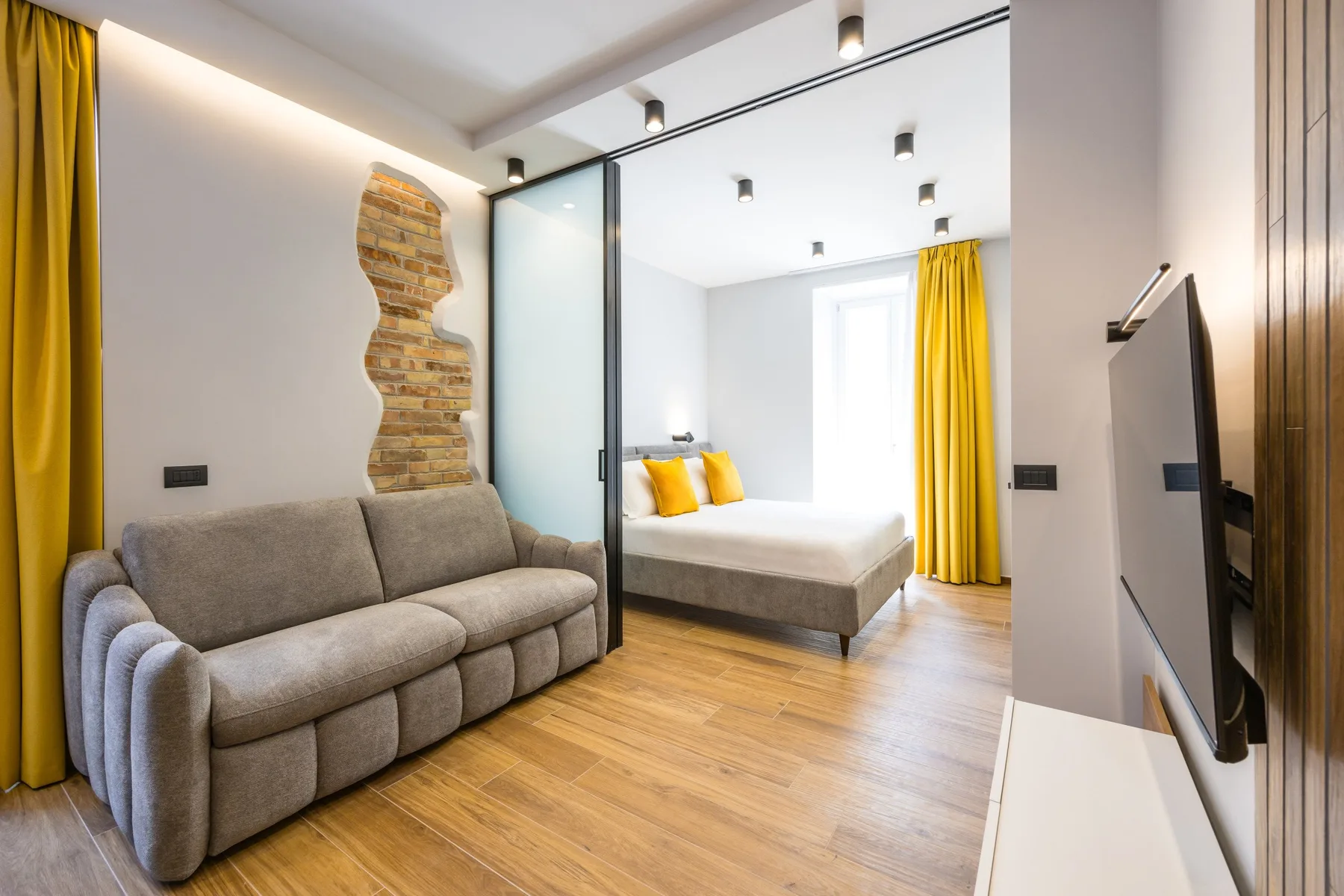 A bright modern room with a gray sofa and a bed behind a glass partition.