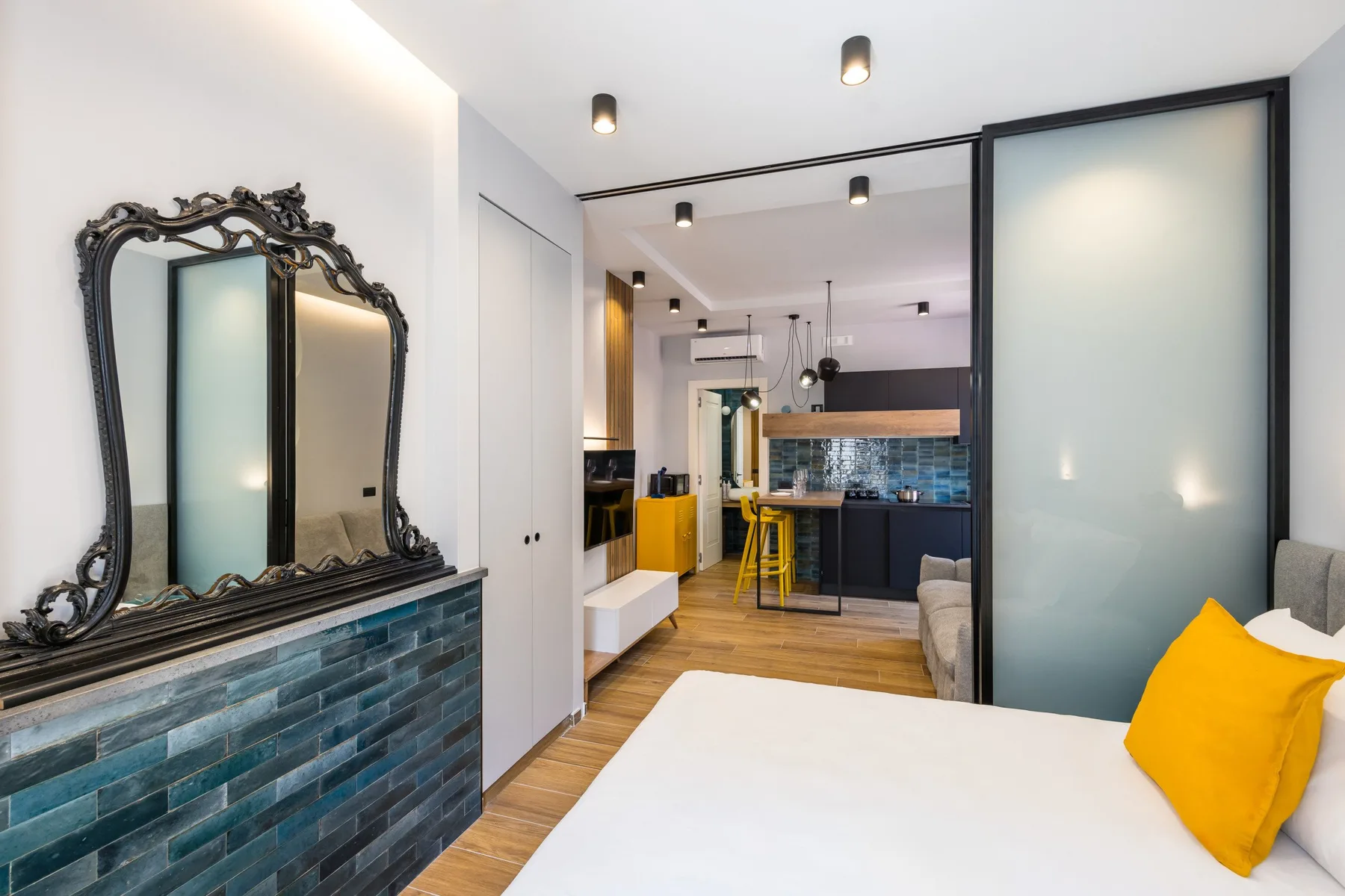 A bedroom opens onto a compact kitchen, with a bed and colorful tiled wall.
