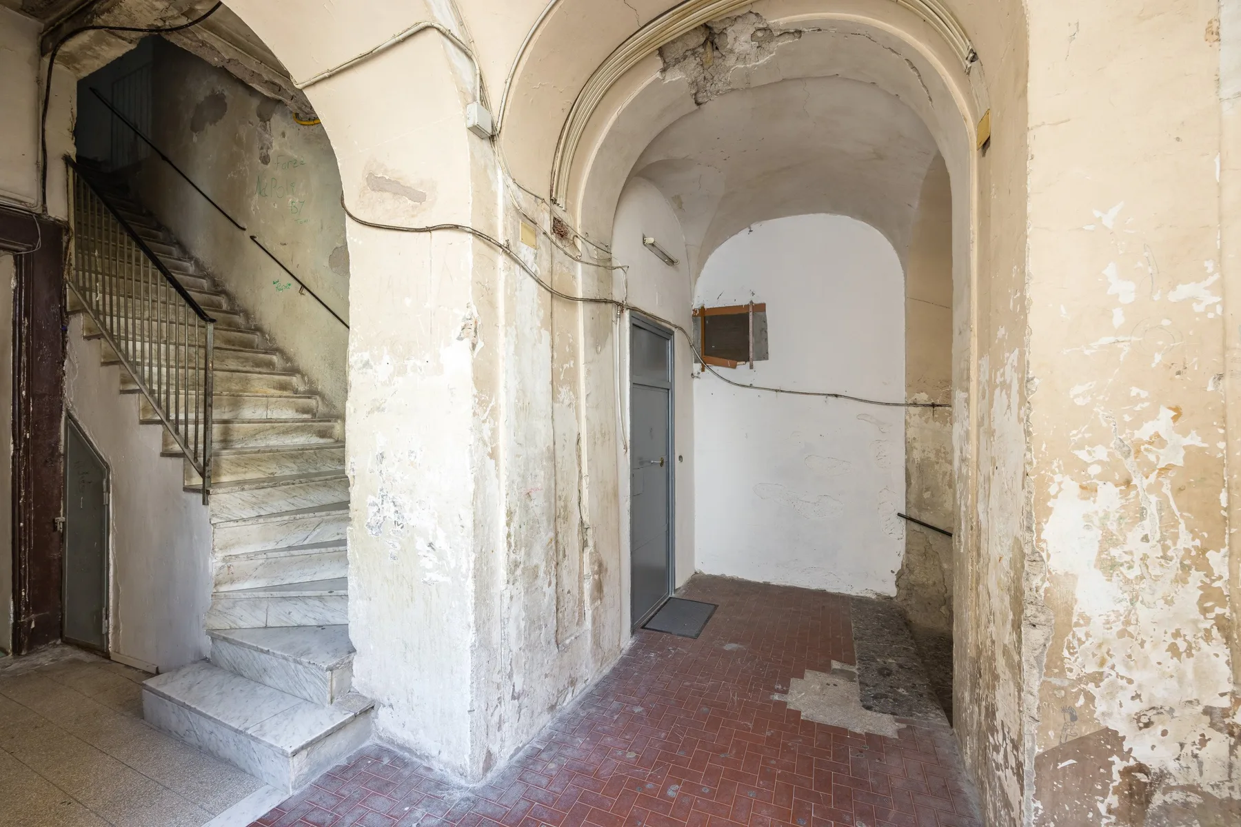 An aged interior hallway with an arched ceiling, tiled floor, and staircase.