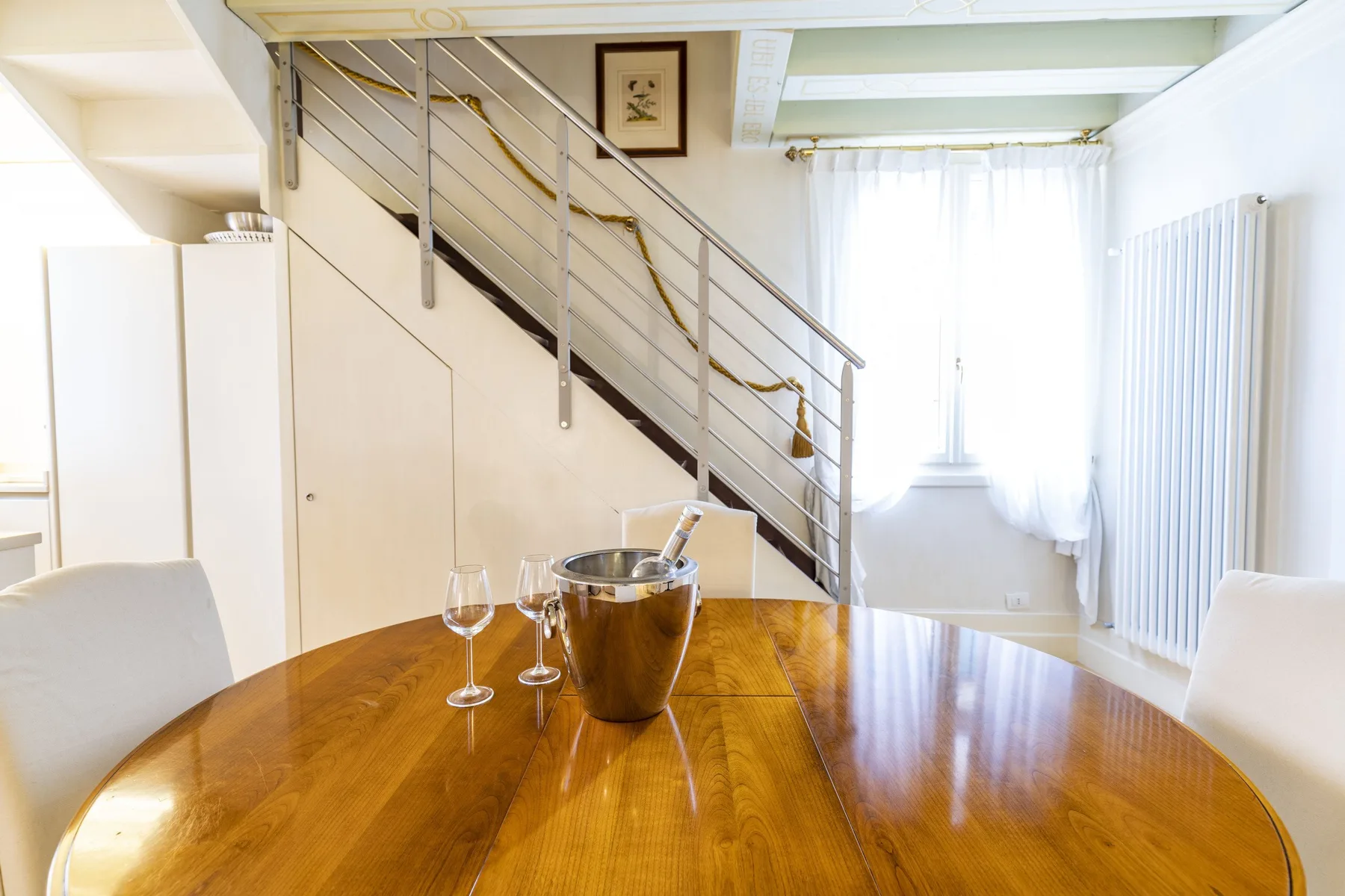 A dining area with a wooden table beneath a staircase and beside a bright window.