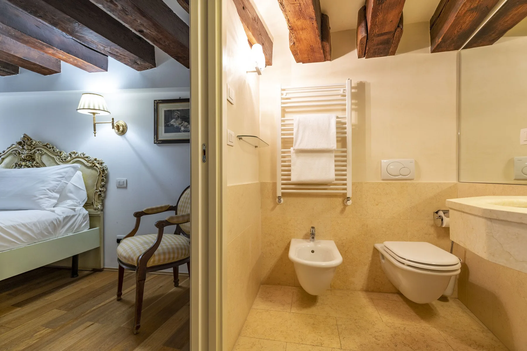 A bathroom with a towel warmer and toilet, beside a bedroom with a bed and chair.