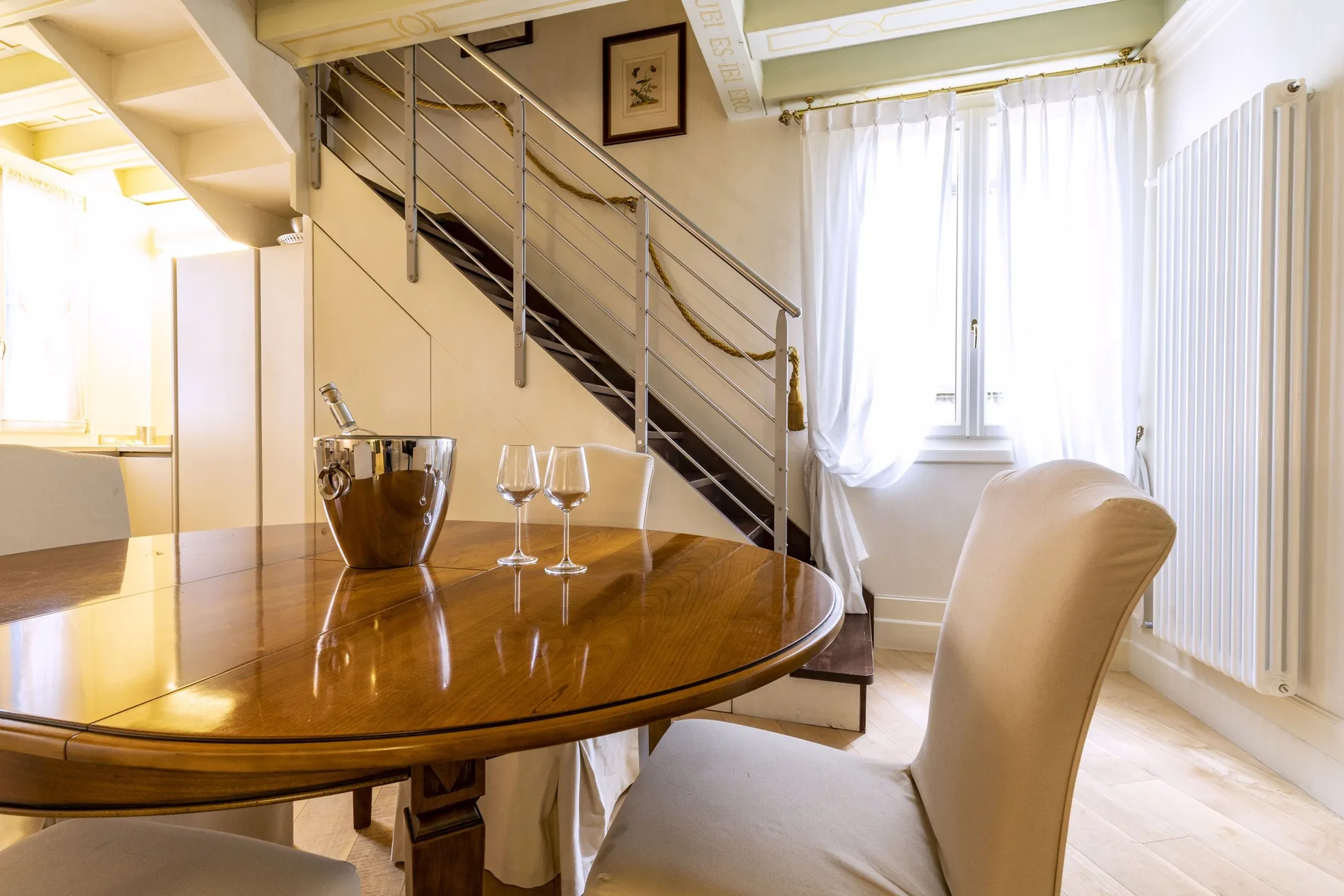 A dining area with a polished oval table beside a staircase and bright window.