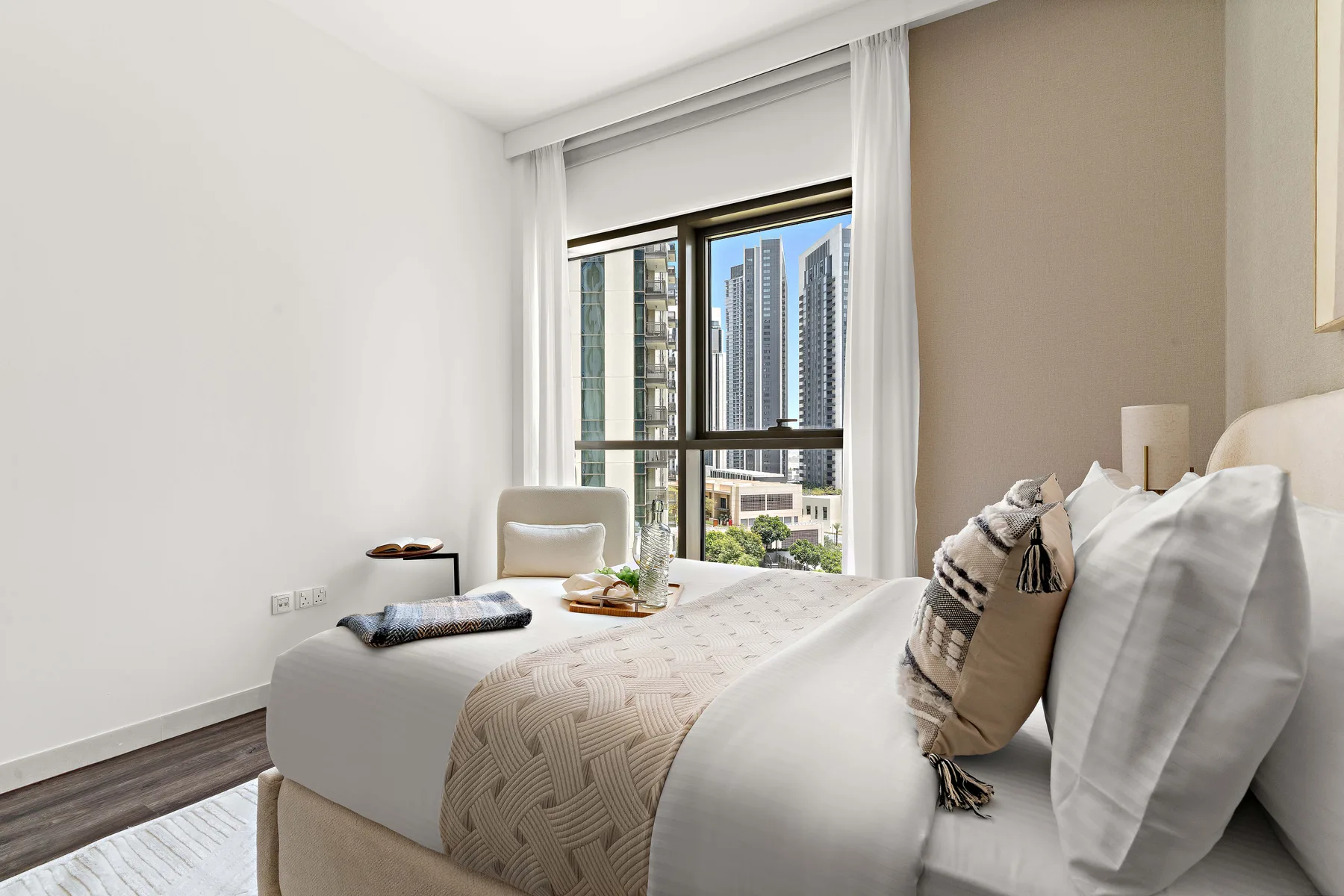 Un dormitorio luminoso con una cama junto a una ventana amplia que da a edificios de la ciudad.