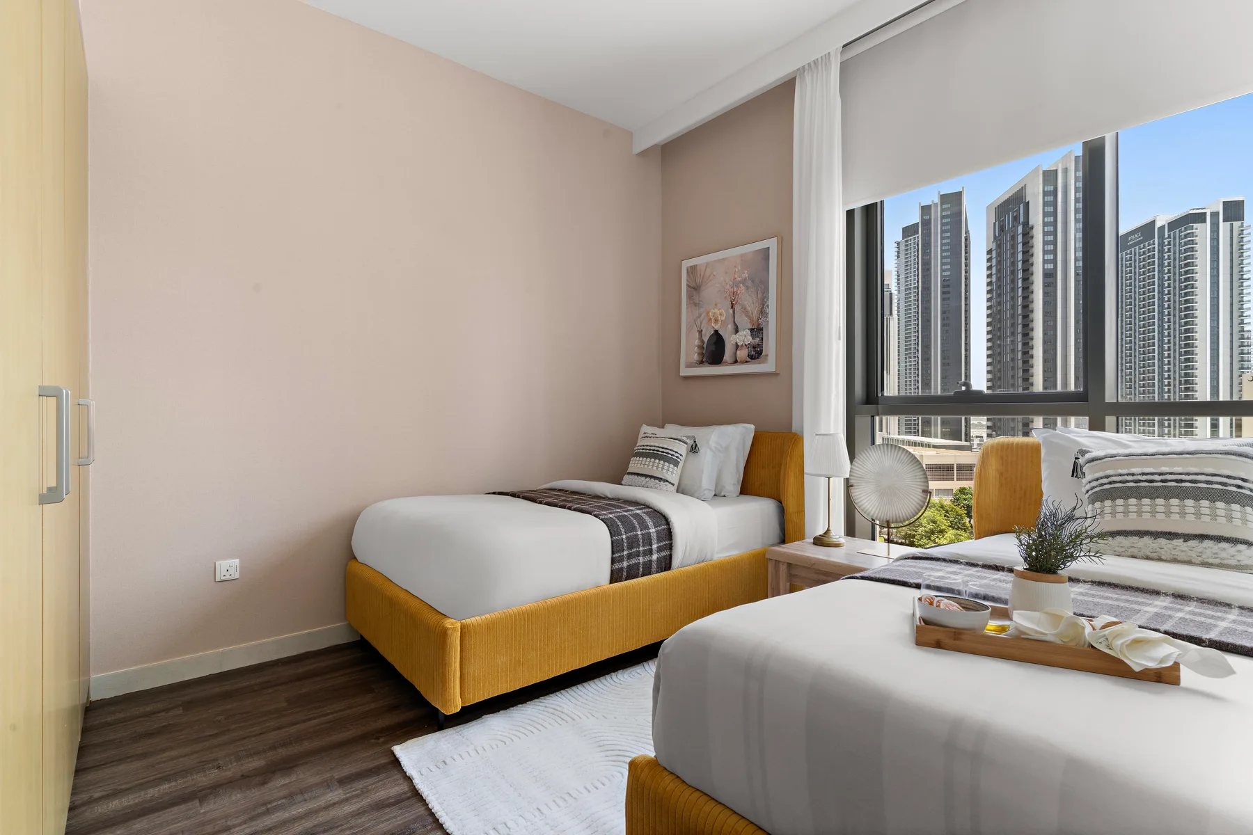 Un dormitorio con dos camas junto a una ventana amplia con vistas a edificios altos.