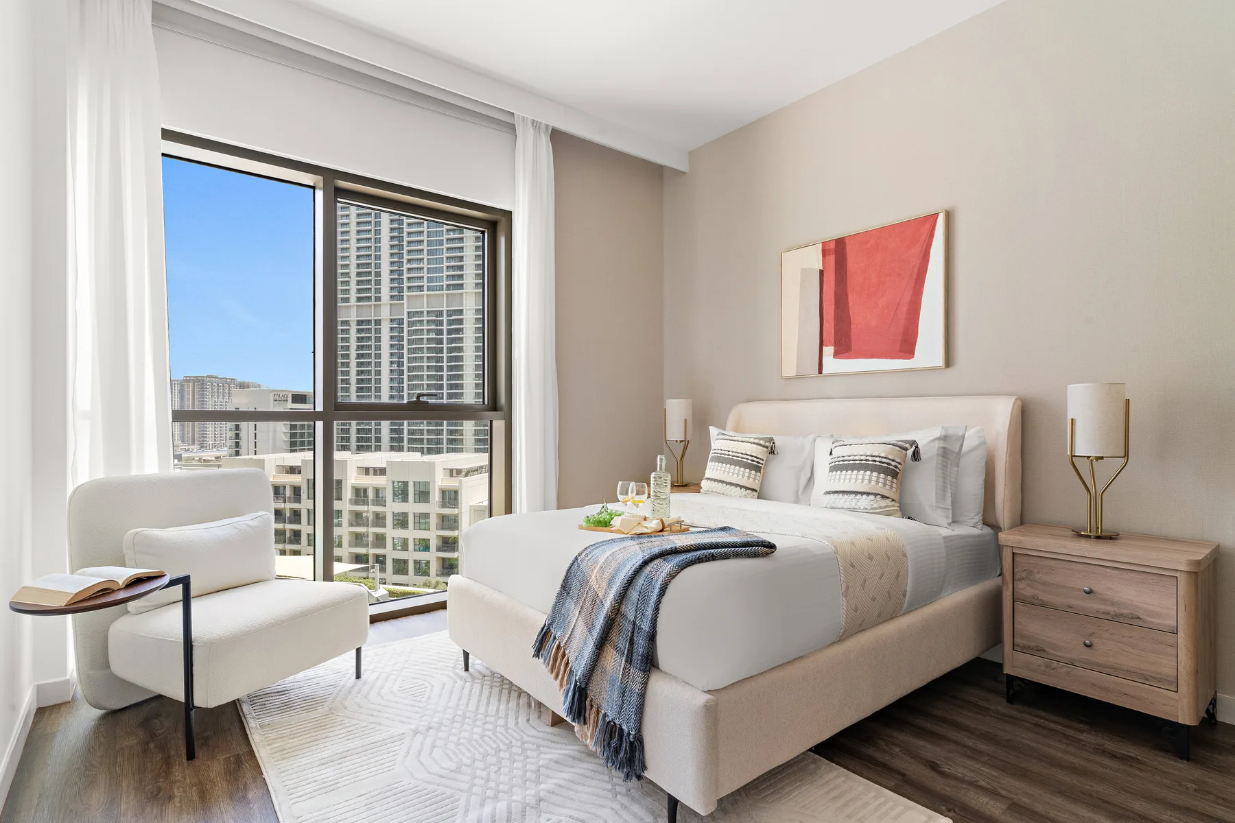 Un dormitorio luminoso con una cama bien tendida y grandes ventanas con vistas a edificios cercanos.