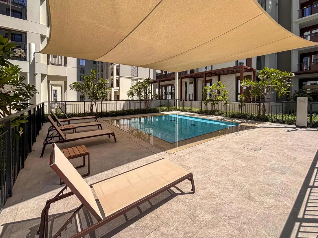 Una piscina exterior está junto a una terraza pavimentada con asientos bajo una cubierta de sombra.