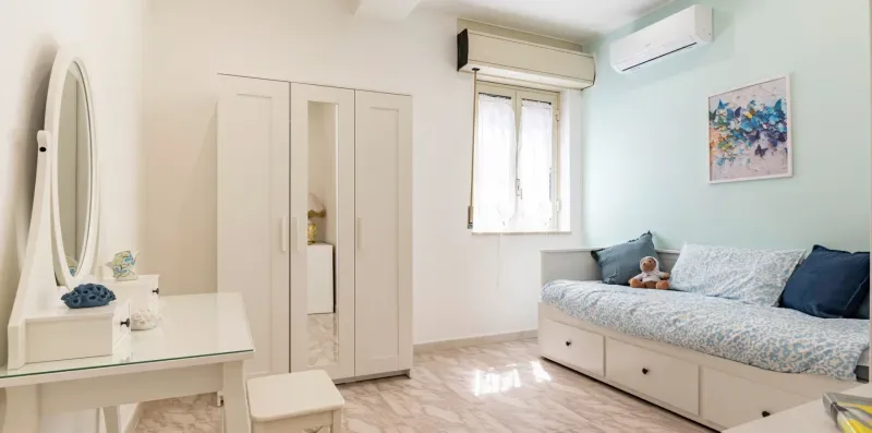 Un dormitorio luminoso con cama, armario y tocador pequeño.