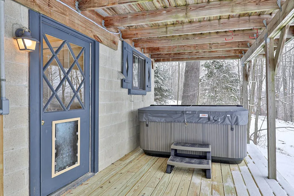 A covered outdoor deck with a hot tub and snowy trees beyond.