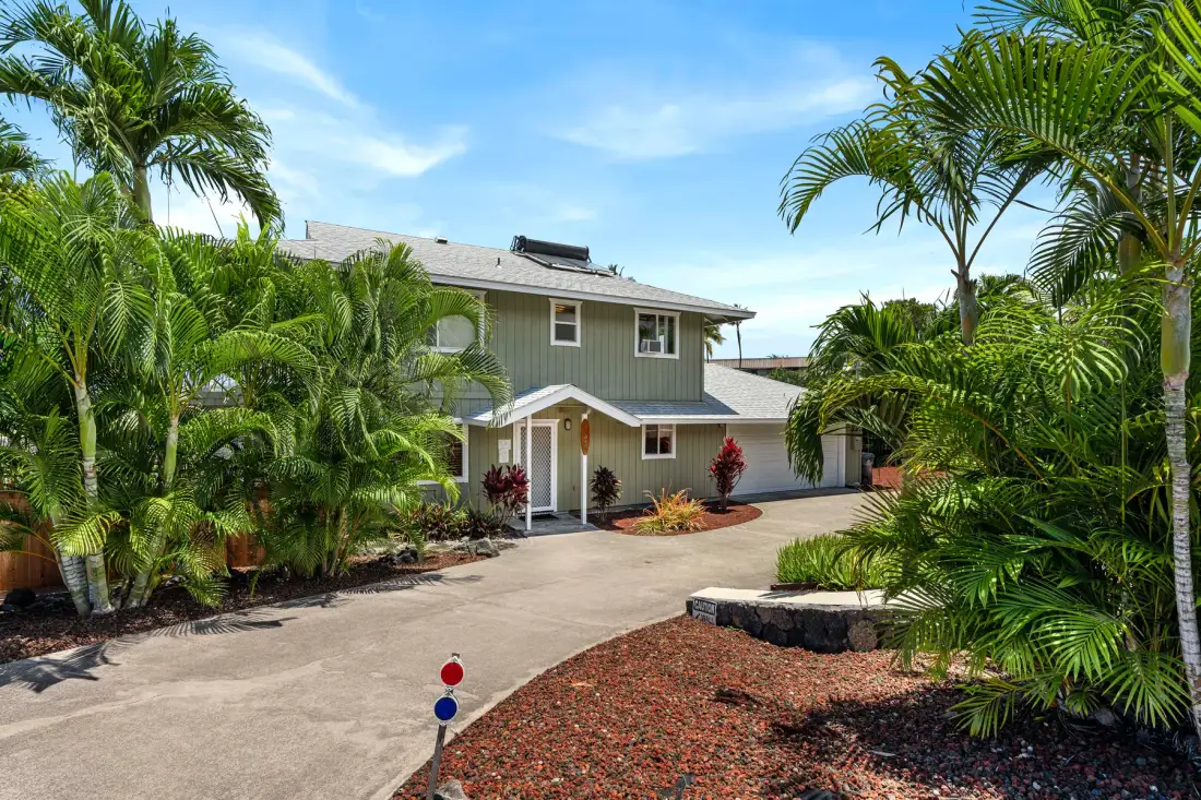 Una casa de dos pisos con entrada pavimentada y vegetación tropical.