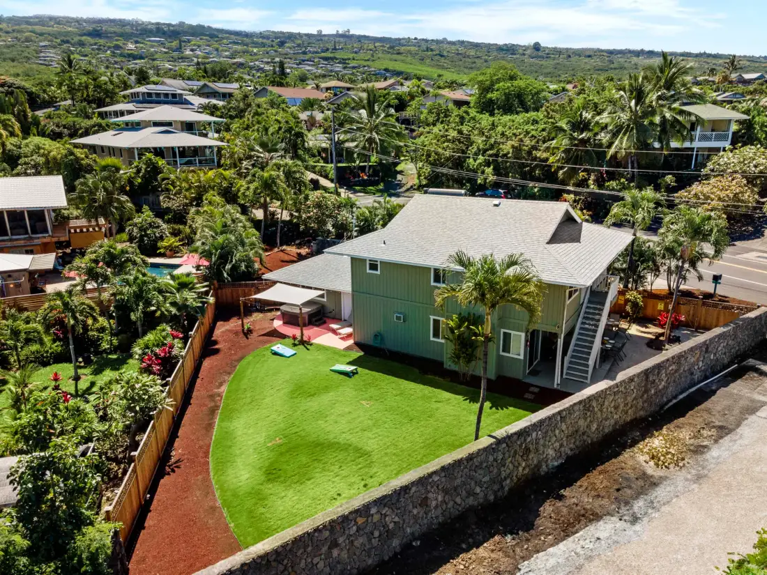 Vista aérea de una casa verde rodeada de jardines tropicales y viviendas vecinas.