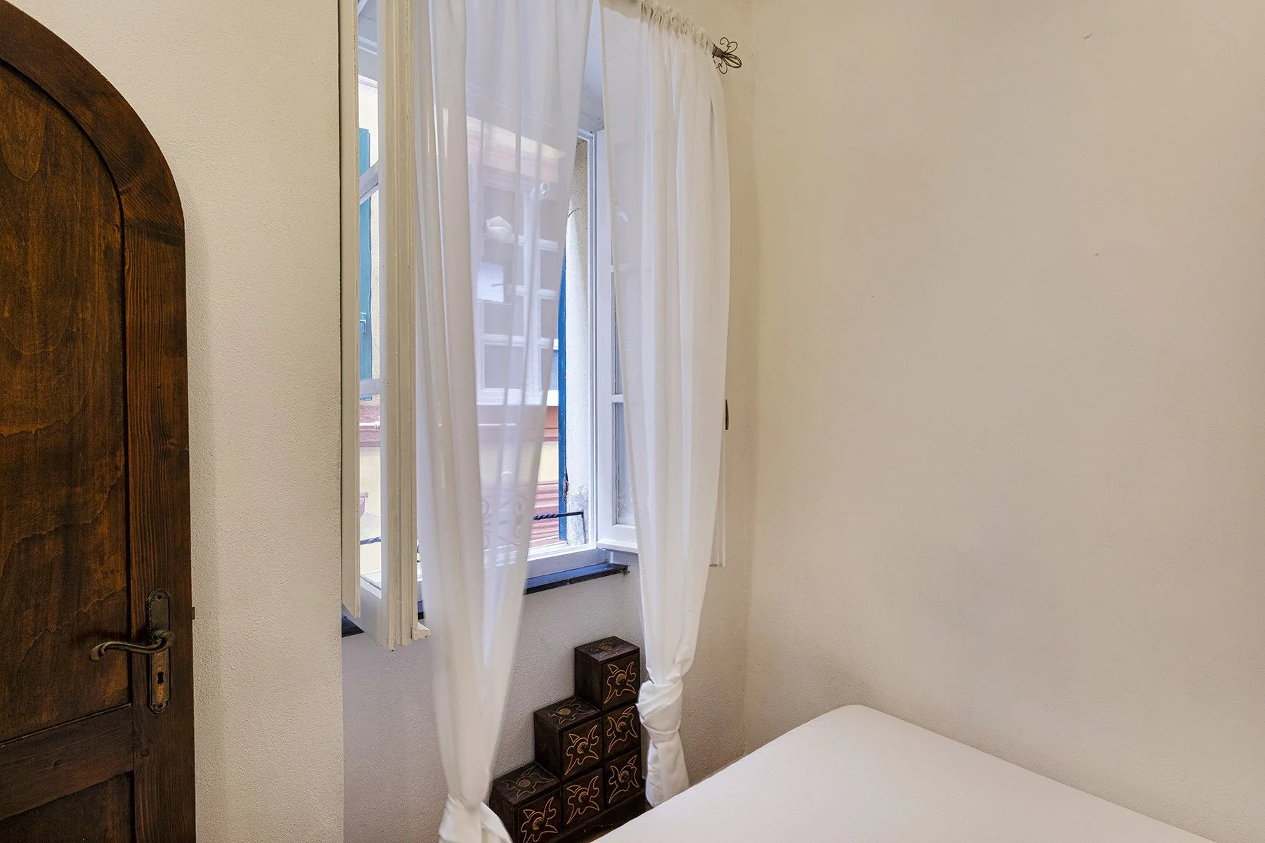 A bedroom with a curtained window beside a wooden door and the edge of a bed.