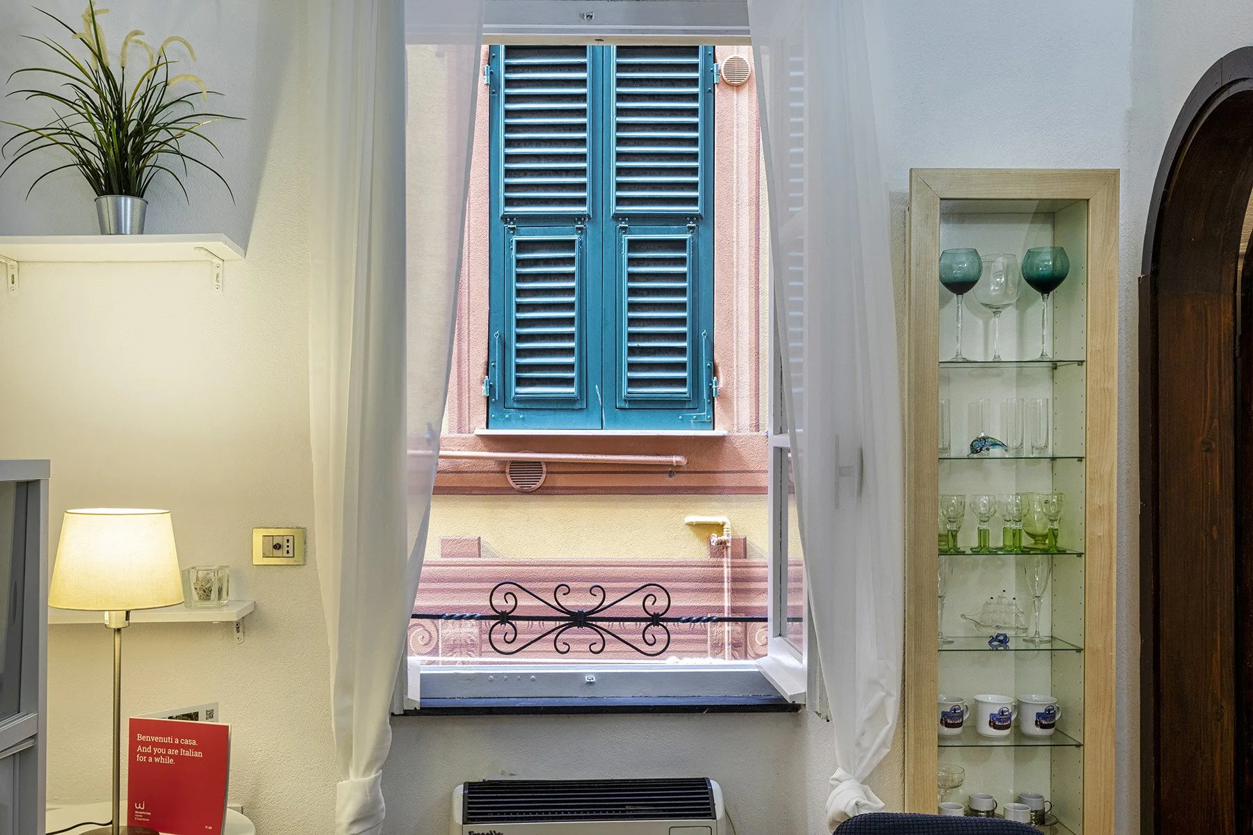 A bright room with an open window overlooking blue shutters and a nearby building.