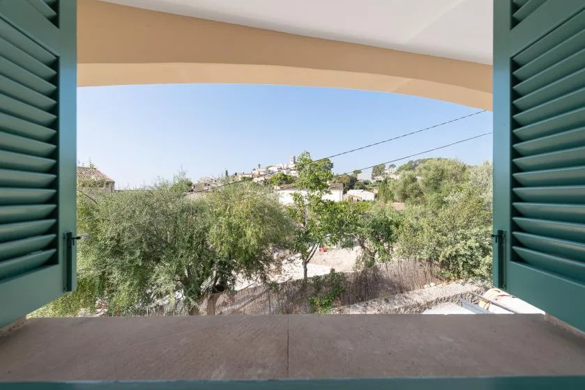 Una ventana abierta con persianas verdes da a árboles y casas cercanas.