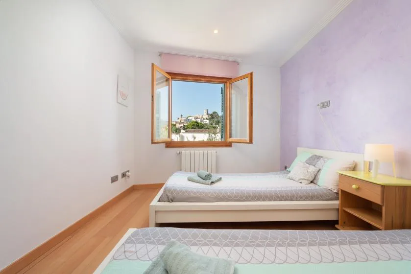 Un dormitorio luminoso con camas y una ventana abierta con vistas a edificios cercanos.