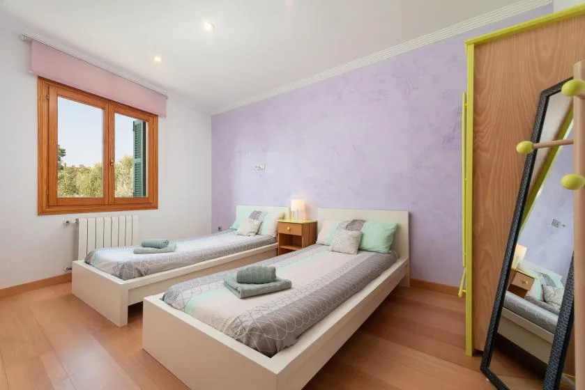 Un dormitorio con dos camas, paredes pastel y una ventana con vistas a vegetación.