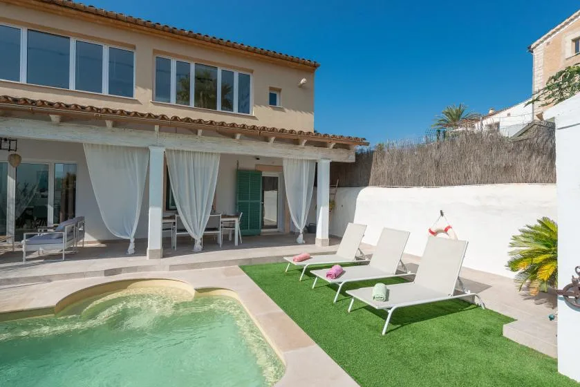 Una zona de piscina al aire libre tiene asientos reclinables sobre césped artificial junto a una casa.