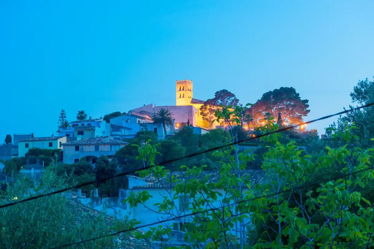 Un pueblo en una ladera al anochecer, con una torre iluminada sobre los edificios cercanos.