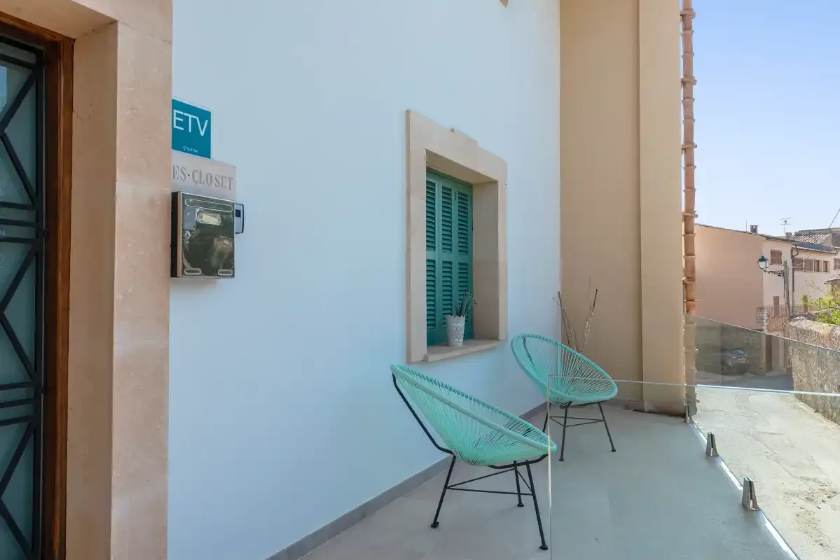 Una pequeña terraza con dos sillas turquesas junto a una pared blanca y una ventana.