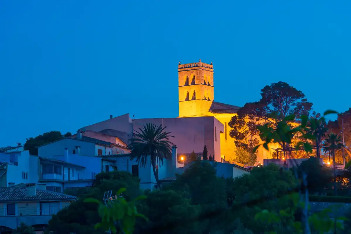 Una torre de iglesia iluminada se alza sobre edificios y árboles al anochecer.