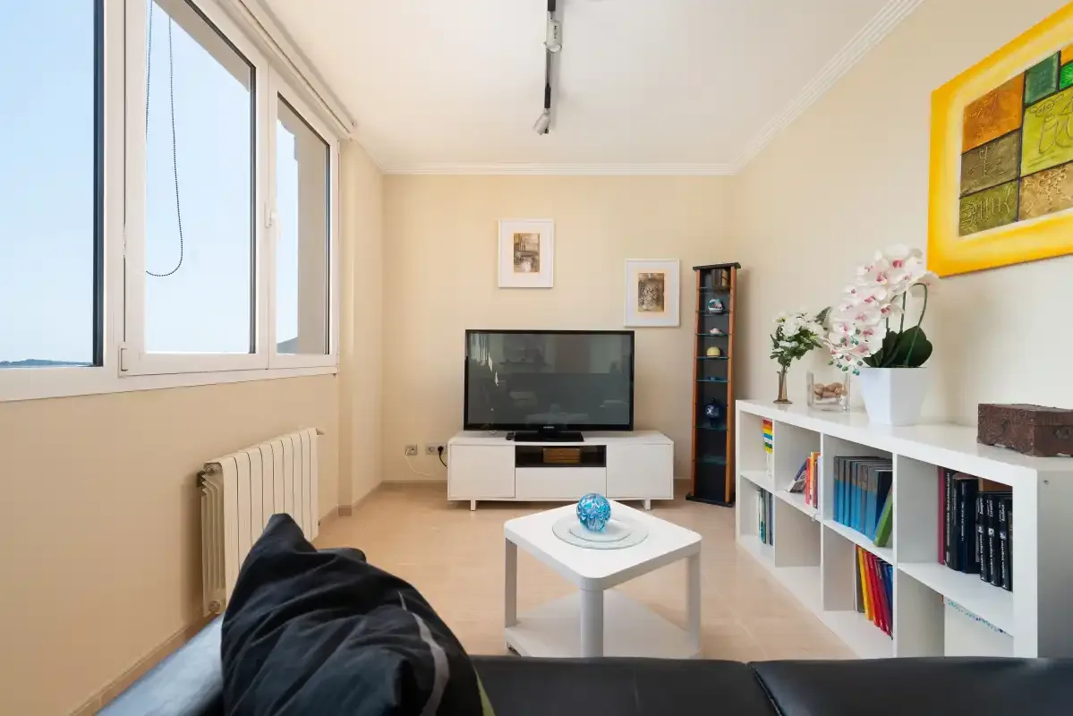 Una sala de estar luminosa con ventanas grandes, un televisor y una mesa baja.