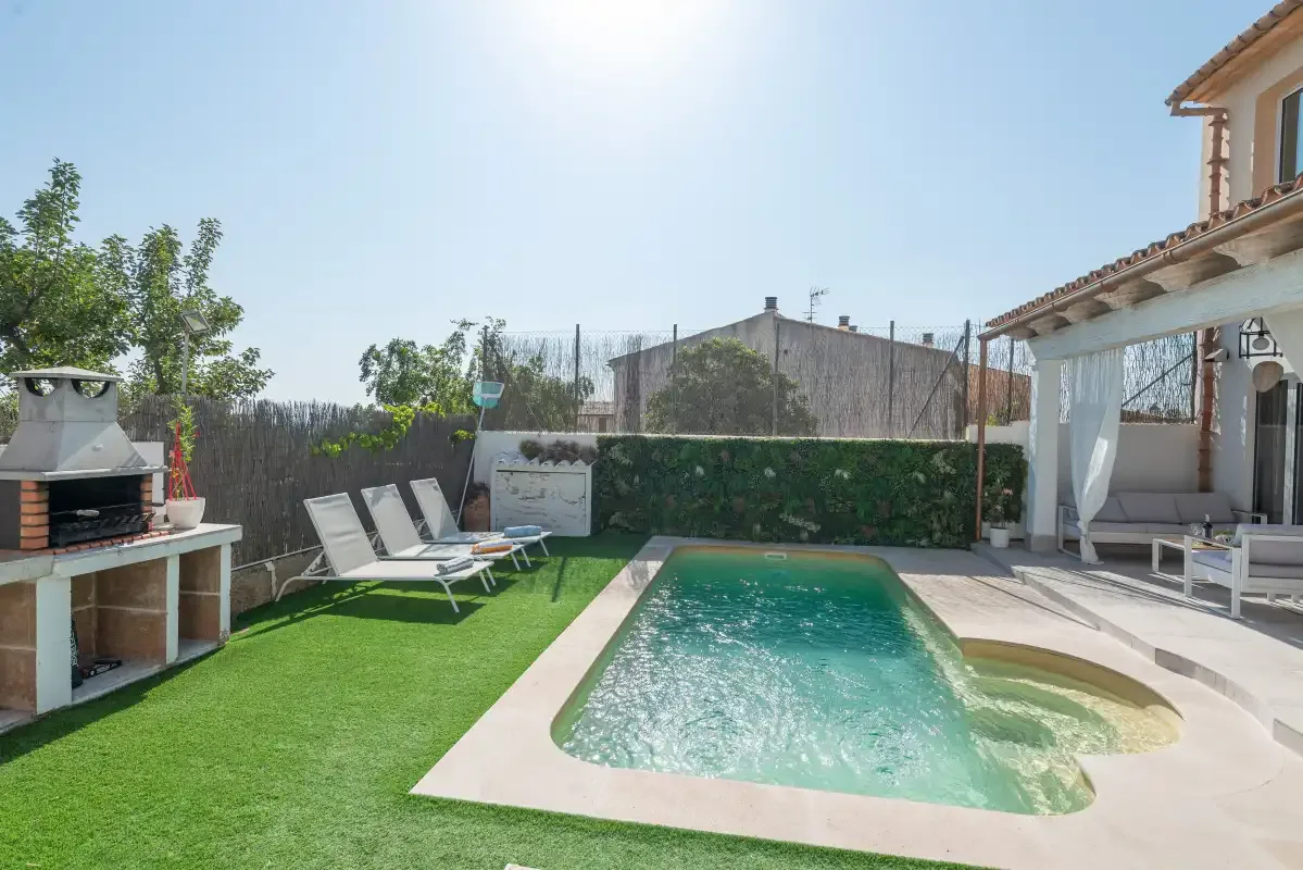 Zona de piscina exterior con tumbonas, césped artificial y un patio cubierto.