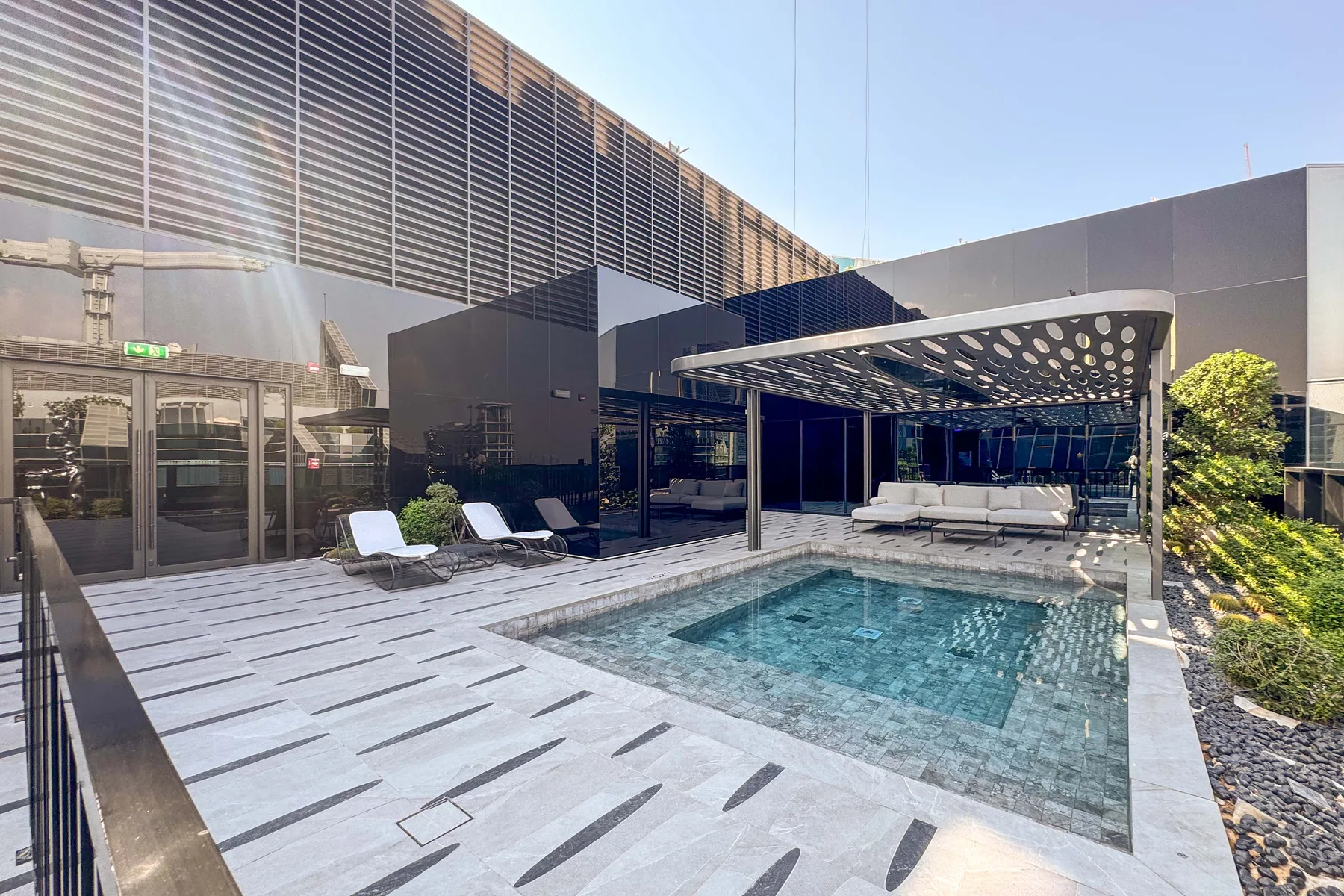 Una piscina exterior junto a un patio pavimentado y una zona de descanso con sombra.