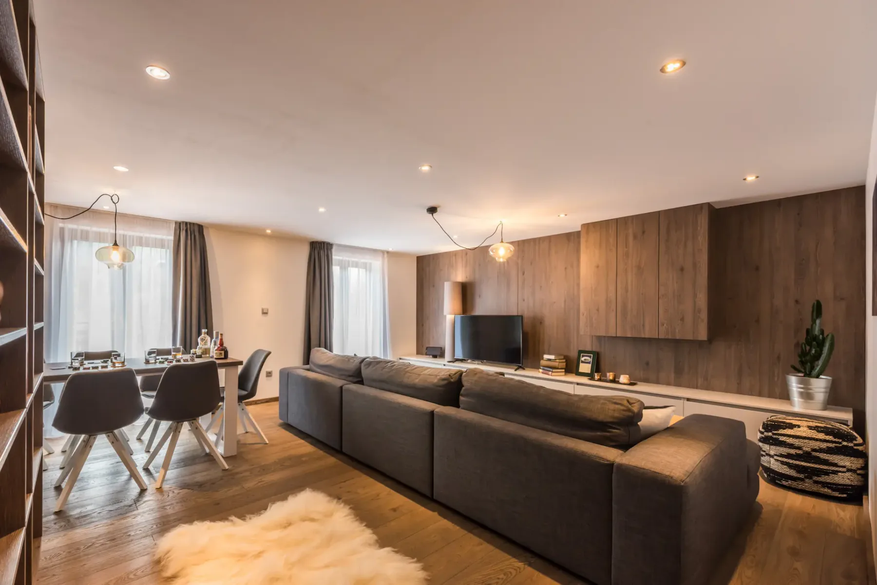 Un salón moderno con asientos frente a un televisor y una zona de comedor junto a las ventanas.