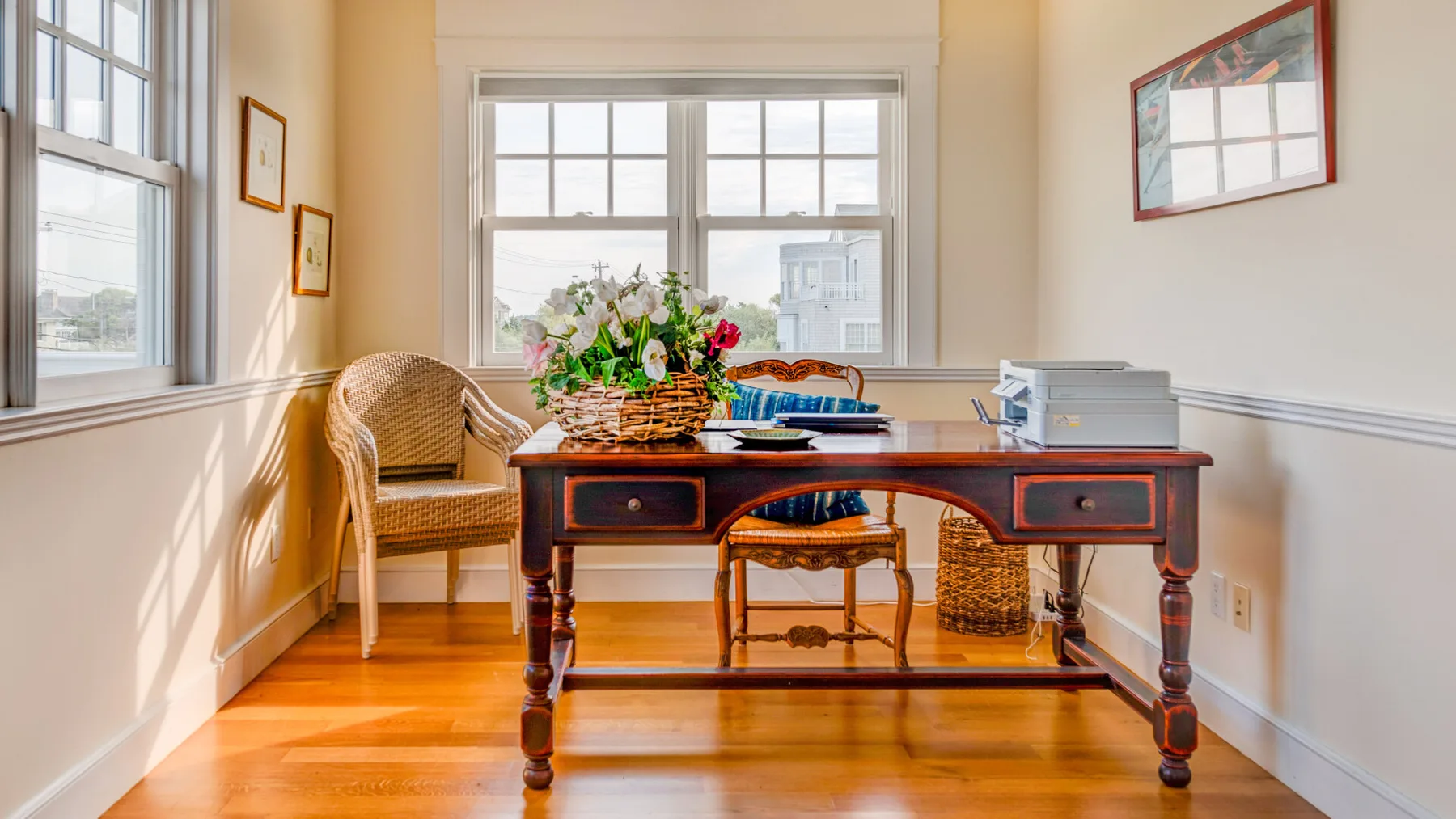 Una habitación luminosa con un escritorio de madera, asientos tejidos y ventanas grandes.