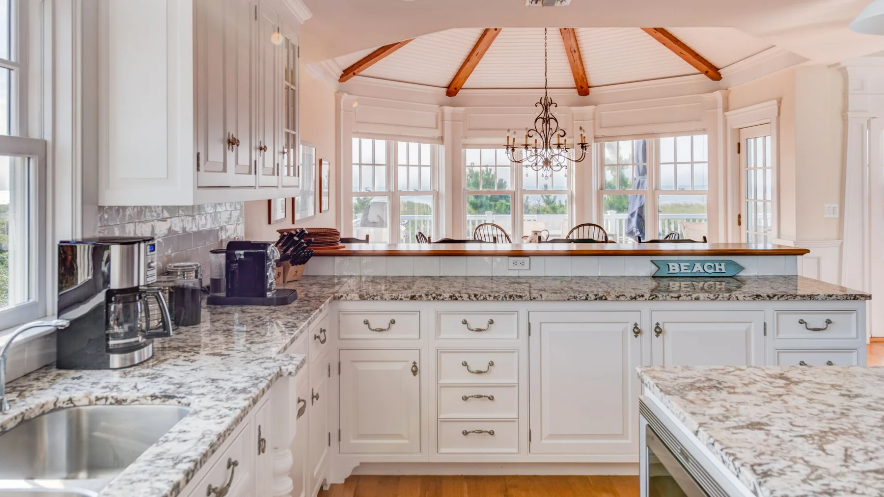 Una cocina luminosa con gabinetes blancos, encimeras de piedra y grandes ventanas.