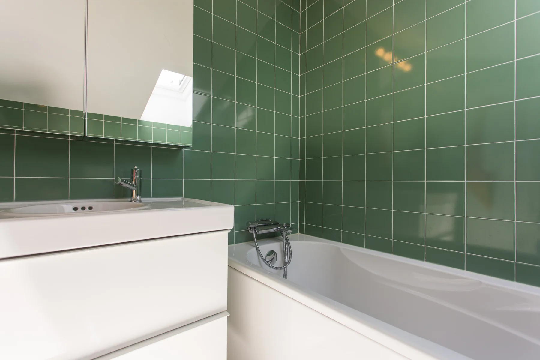 Un baño con un mueble lavabo blanco y una bañera junto a paredes de azulejos verdes.